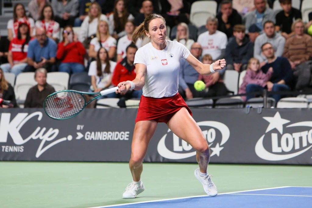Katarzyna Kawa w trakcie turnieju Billie Jean King Cup w Gorzowie Wielkopolskim Tenisistka w białej koszulce z polskim godłem i czerwonej spódniczce odbija piłkę rakietą podczas meczu, w tle widownia skupiona na rozgrywce, na korcie widoczne reklamy sponsorów.