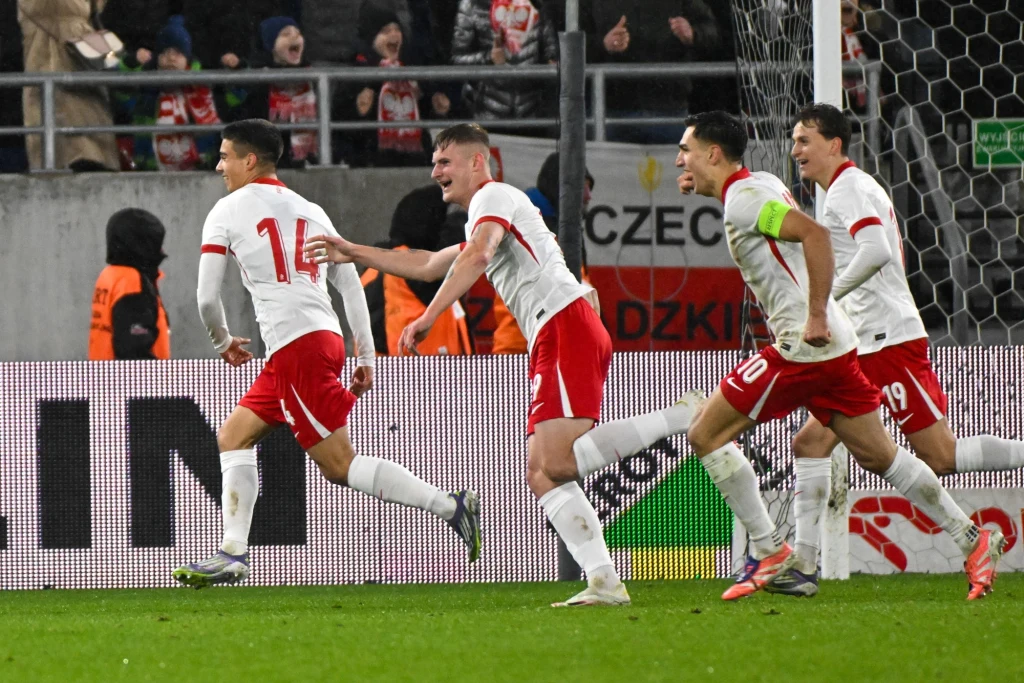 Reprezentacja Polski U-21 Grupa piłkarzy w biało-czerwonych strojach biegnie po murawie stadionu, świętując zdobycie gola, w tle widoczna publiczność oraz siatka bramki.