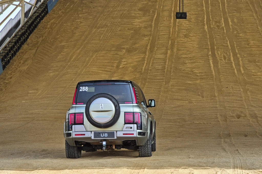 Yangwang U8 dzięki ogromnej mocy i czterem elektrycznym silnikom z łatwością radzi sobie na stromej wydmie Samochód terenowy z napisem 288 na tylnej szybie stoi u podnóża stromego, piaszczystego podjazdu. W tle widoczne ślady opon, a po bokach elementy ogrodzenia toru.