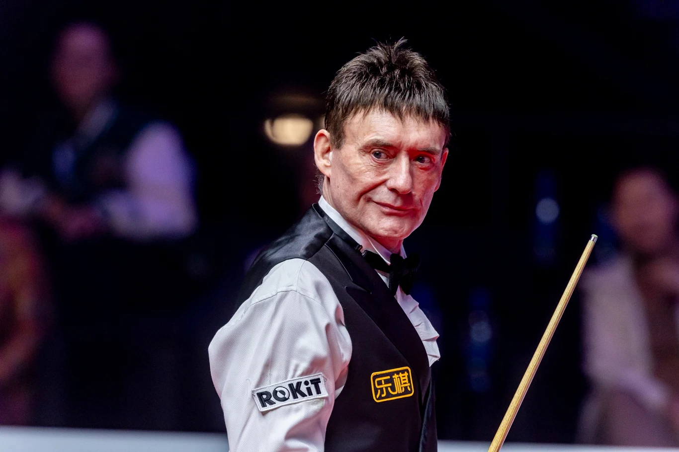 Jimmy White Mężczyzna ubrany w elegancką kamizelkę oraz koszulę, stojący z kijem bilardowym na arenie snookera, w tle nieostre postacie oraz ciemne otoczenie wskazujące na profesjonalny turniej.