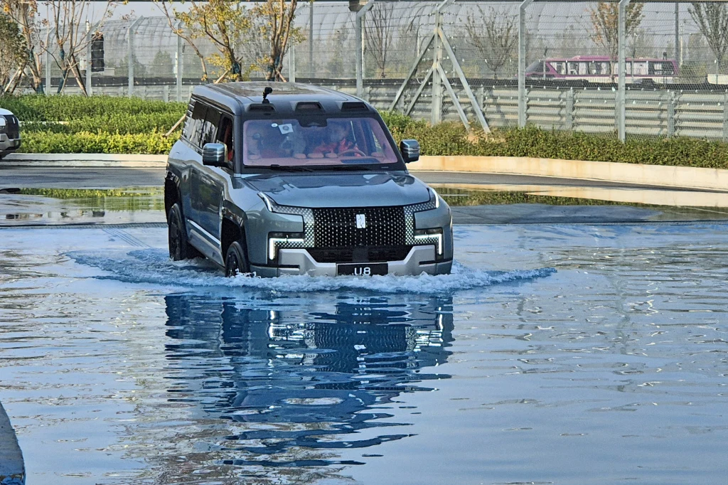 Yangwang U8 posiada także funkcję pływania Luksusowy czarny SUV przeprawiający się przez płytki zbiornik wodny, otoczony nowoczesnym ogrodzeniem i niskimi krzewami, w tle widoczne drzewa i elementy infrastruktury.