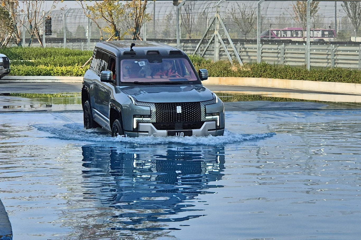 Yangwang U8 posiada także funkcję pływania Luksusowy czarny SUV przeprawiający się przez płytki zbiornik wodny, otoczony nowoczesnym ogrodzeniem i niskimi krzewami, w tle widoczne drzewa i elementy infrastruktury.