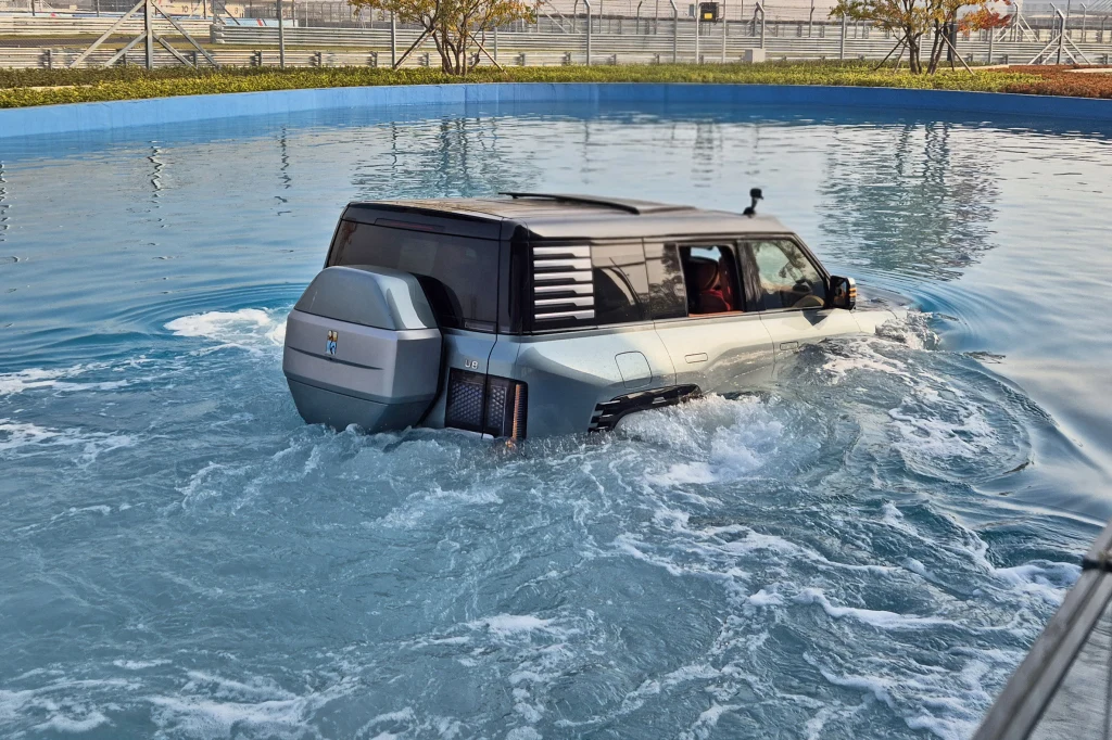 Podczas pływania przód Yangwanga U8 wyraźnie nurkuje, ale nie jest to nic groźnego Samochód terenowy częściowo zanurzony w wodzie, płynący przez specjalny basen testowy otoczony niskim murem. Wokół widoczna jest infrastruktura przemysłowa oraz elementy ogrodzenia.