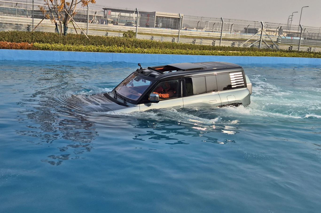 Podczas pływania szklany dach automatycznie się otwiera, aby droga do ewentualnej ucieczki zawsze była gotowa Samochód terenowy poruszający się po powierzchni wody, częściowo zanurzony, otoczony niebieskim basenem lub kanałem, w tle ogrodzenie i zabudowania przemysłowe.
