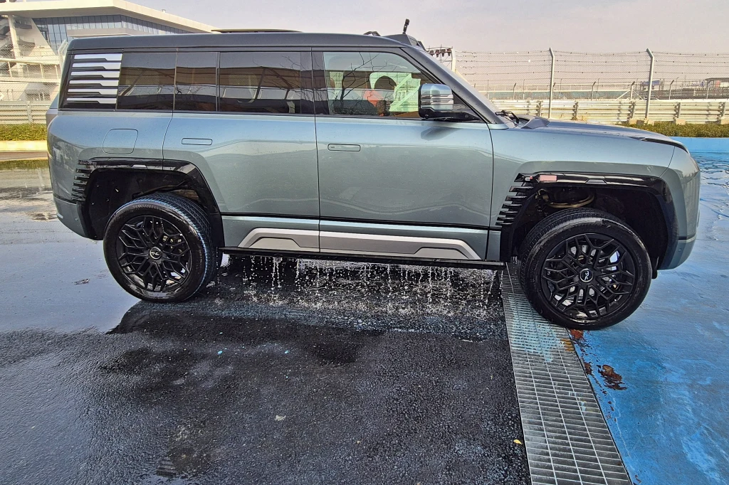 Po wyjechaniu z basenu z U8 wylewa się sporo wody, ale nie dostaje się ona do newralgicznych elementów pojazdu ani do wnętrza Luksusowy, nowoczesny SUV o dużym prześwicie unoszący się nad kratką ściekową, woda wypływa spod pojazdu, a w tle widoczny jest tor wyścigowy otoczony ogrodzeniem.