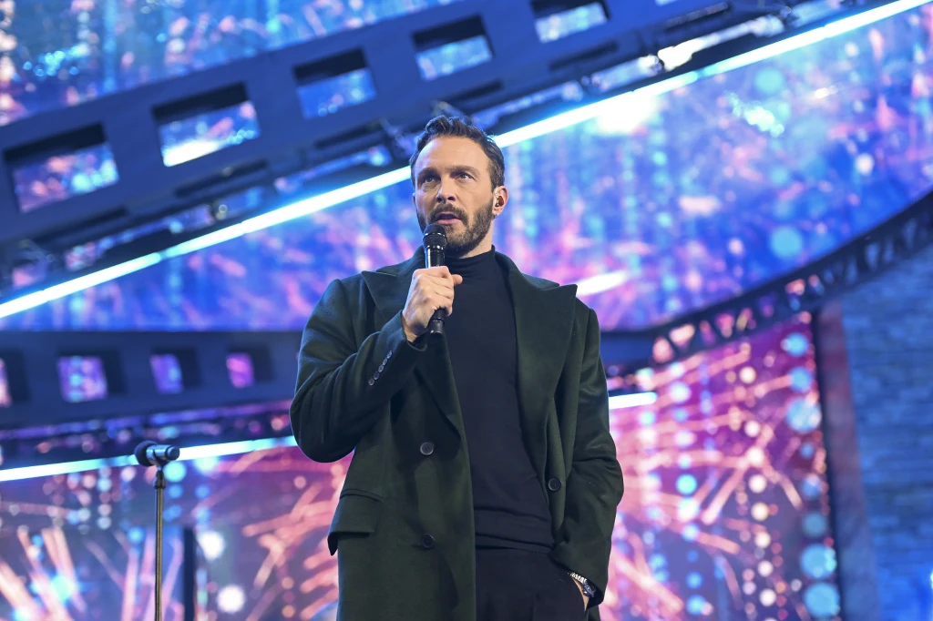 Sławek Uniatowski miał wypadek samochodowy w drodze na koncert w Zamościu Mężczyzna w ciemnym płaszczu trzyma mikrofon na scenie, za nim widać barwne światła i dekoracje sceniczne w formie taśmy filmowej.