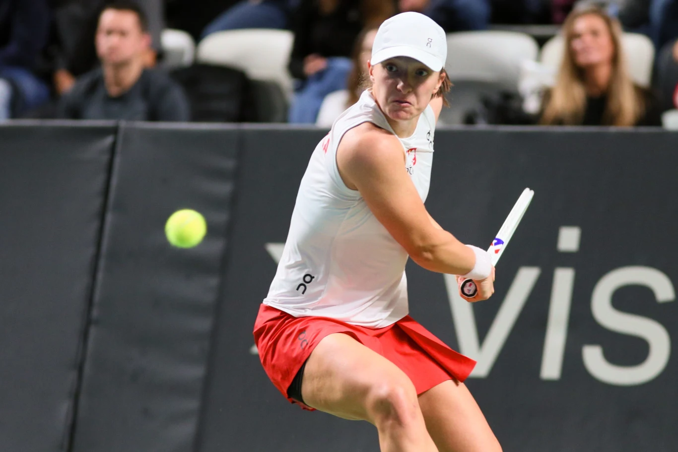 Tenisistka uderzająca piłkę rakietą podczas meczu na korcie, ubrana w białą koszulkę, czerwoną spódniczkę i czapkę, w tle widoczni kibice.