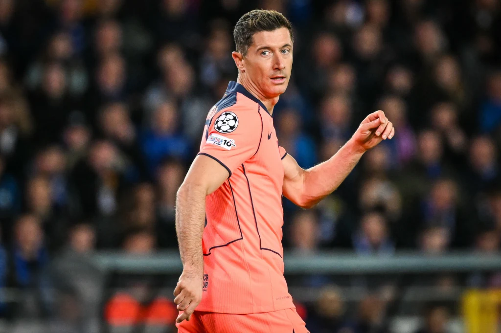 Robert Lewandowski Piłkarz ubrany w pomarańczowy strój sportowy patrzy przez ramię podczas meczu, widoczne logo Ligi Mistrzów na rękawie i tłum kibiców w tle.