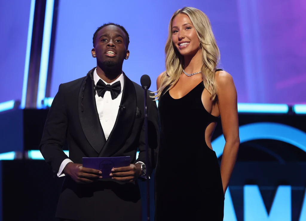 Dwójka prowadzących na scenie podczas oficjalnego wydarzenia, mężczyzna w czarnym smokingu z muchą i kobieta w długiej, czarnej sukni, stoją razem przy mikrofonie na tle kolorowego oświetlenia.