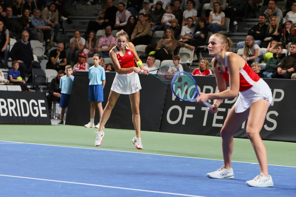 Linda Klimovicova i Martyna Kubka w meczu Billie Jean King Cup Dwie zawodniczki w czerwonych koszulkach i białych spódniczkach na korcie tenisowym w trakcie deblowego meczu, przygotowane do odbioru piłki, za nimi widoczna publiczność oraz sędziowie i dzieci do podawania piłek.