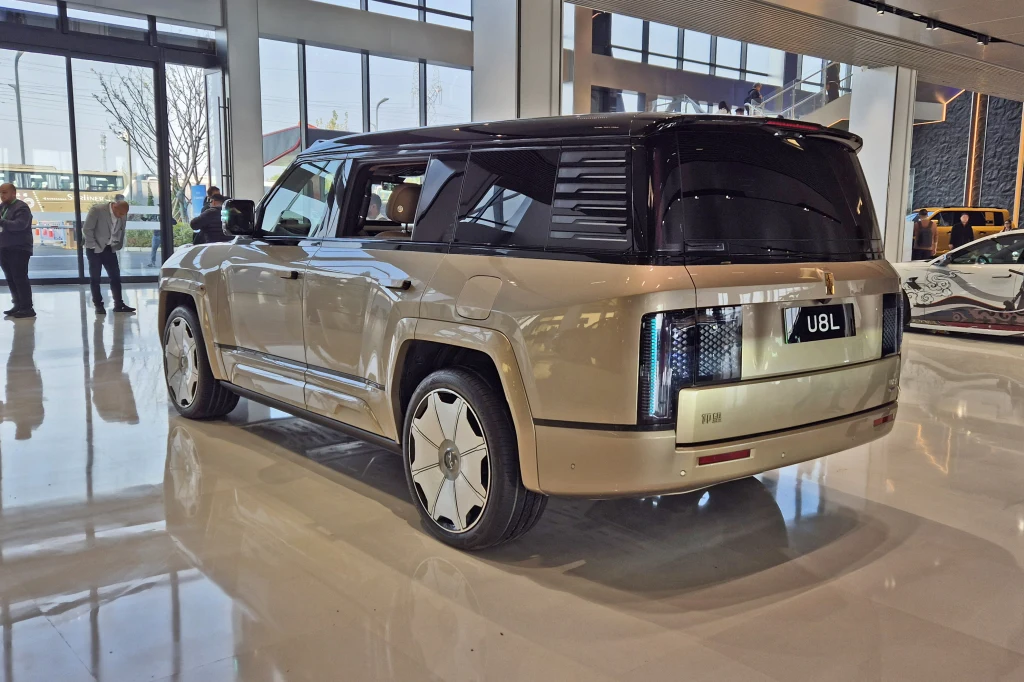 Yangwang U8L, czyli przedłużona wersja z powodzeniem mogąca zastąpić reprezentacyjną limuzynę Luksusowy SUV w jasnym kolorze prezentowany w nowoczesnym salonie samochodowym, wyróżniający się futurystycznym designem, unikalnymi tylnymi światłami oraz dużymi, ozdobnymi felgami. W tle widać inne pojazdy oraz osoby przyglądające się ekspozycji.
