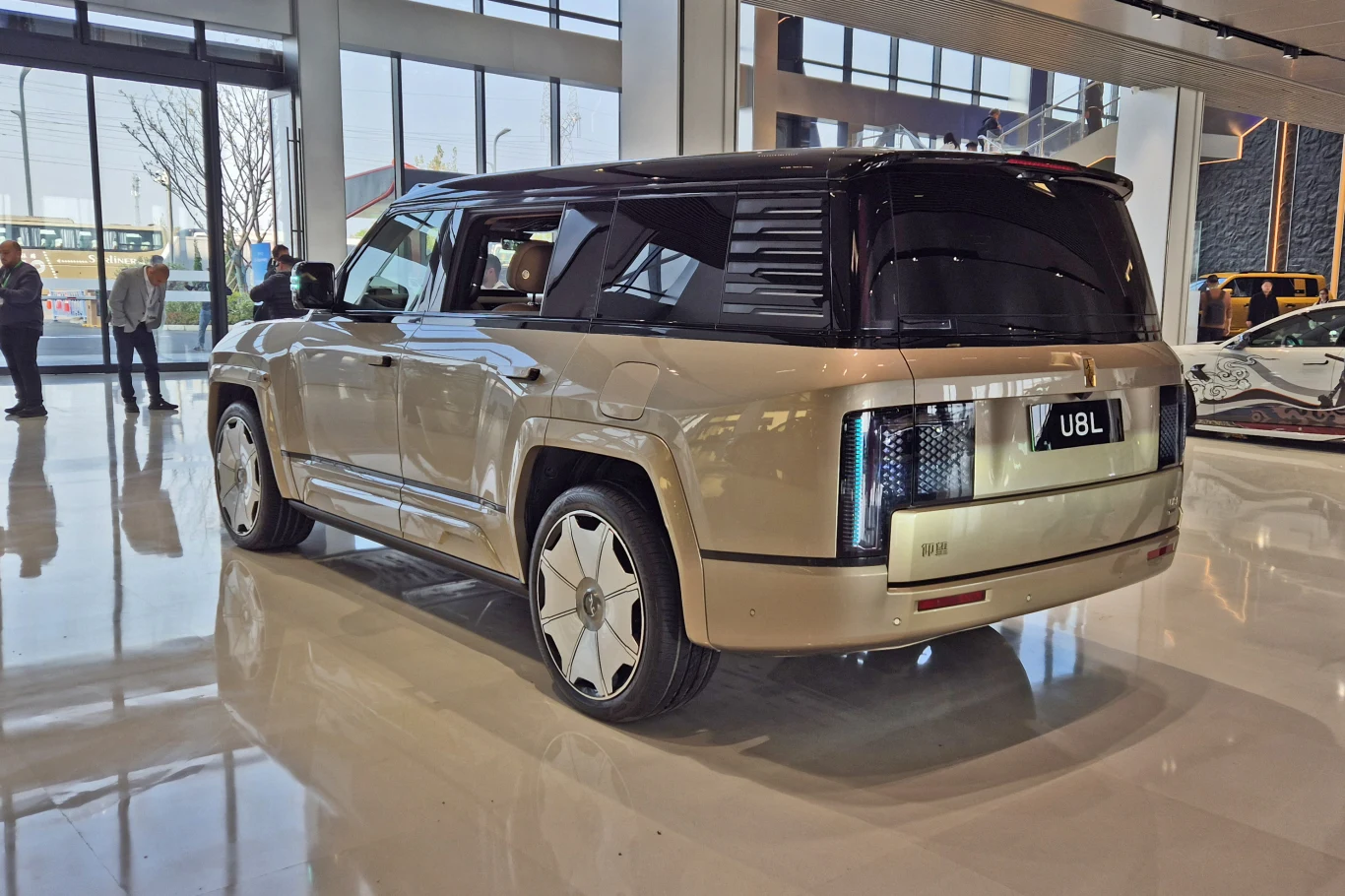 Yangwang U8L, czyli przedłużona wersja z powodzeniem mogąca zastąpić reprezentacyjną limuzynę Luksusowy SUV w jasnym kolorze prezentowany w nowoczesnym salonie samochodowym, wyróżniający się futurystycznym designem, unikalnymi tylnymi światłami oraz dużymi, ozdobnymi felgami. W tle widać inne pojazdy oraz osoby przyglądające się ekspozycji.