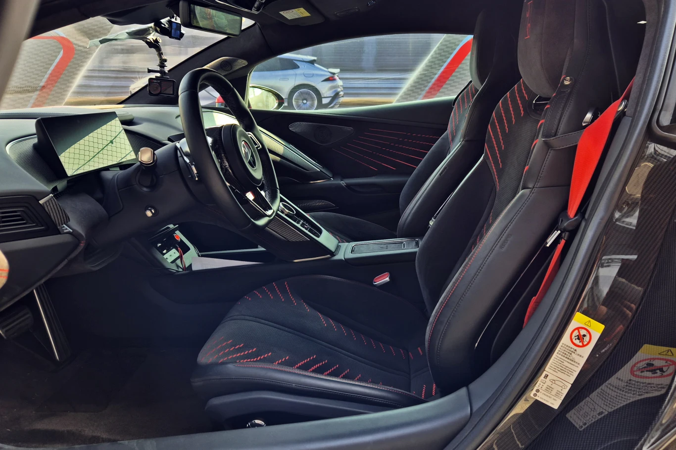 Fotele są niezłe, ale równie dobrze mogłyby trafić do zwykłego samochodu Nowoczesne wnętrze sportowego samochodu z czarnymi fotelami obszytymi czerwonymi nićmi, widoczną kierownicą, wyświetlaczem na desce rozdzielczej i detalami technologii.