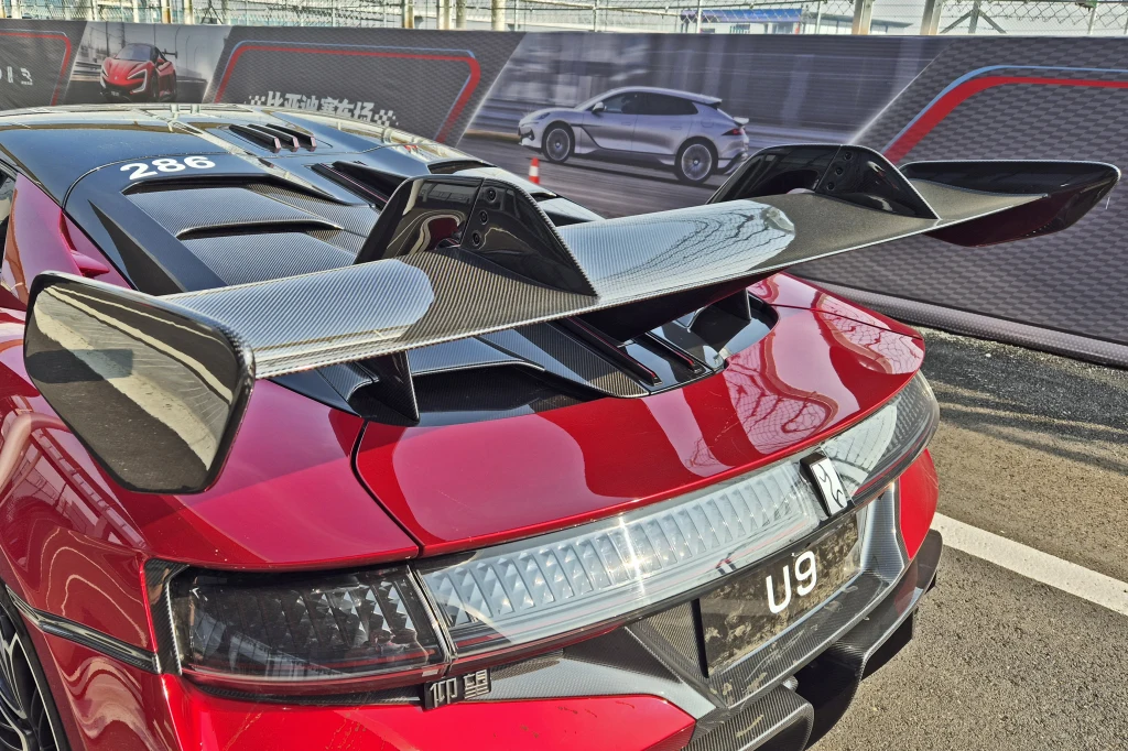 Wielkie karbonowe skrzydło to opcja, zapewniająca dodatkowe 280 kg docisku Czerwony sportowy samochód z widocznym ogromnym tylnym spojlerem z włókna węglowego, szczegółowo ukazany fragment tylnej części, błyszczące nadwozie oraz tablica rejestracyjna z napisem U9, w tle baner z grafiką innego auta.