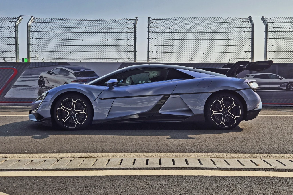 Trudno w to uwierzyć, ale U9 ma niemal 5 m długości Nowoczesny srebrny samochód sportowy ustawiony na torze wyścigowym, otoczony ogrodzeniem, na tle grafiki przedstawiającej inne auta wyścigowe.