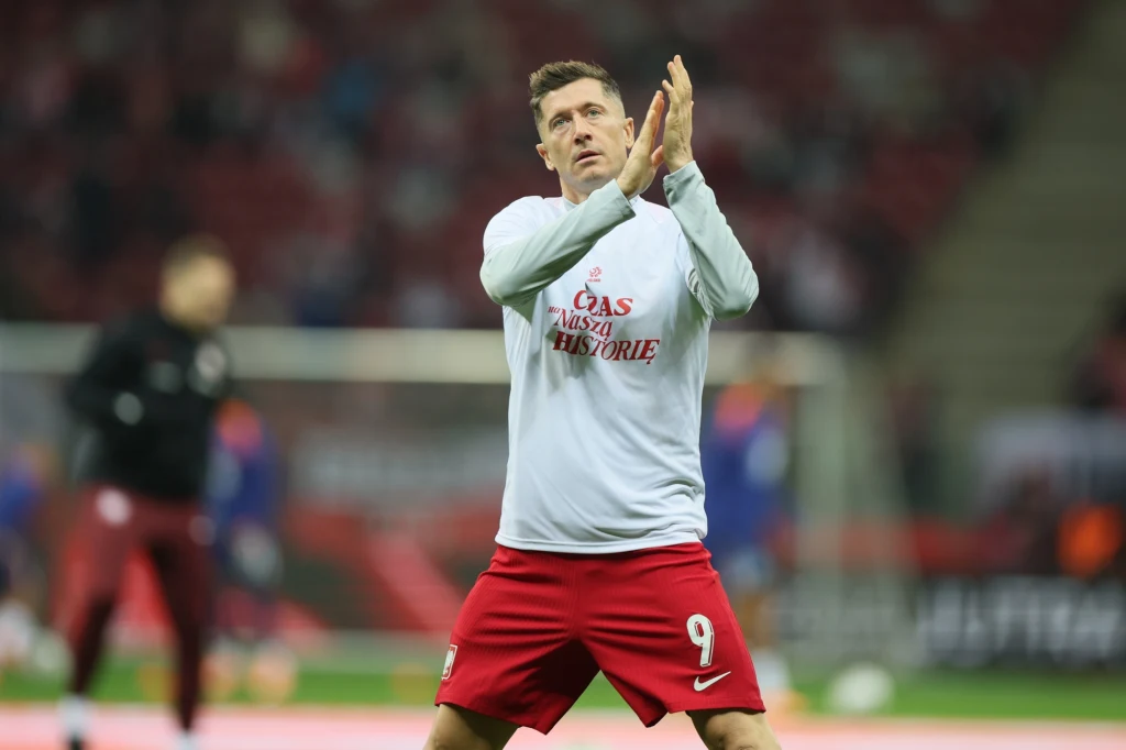 Robert Lewandowski Piłkarz ubrany w białą koszulkę z napisem i czerwone spodenki stoi na boisku stadionowym, wykonując gest oklasków. W tle rozmazane sylwetki innych osób oraz trybuny stadionu.