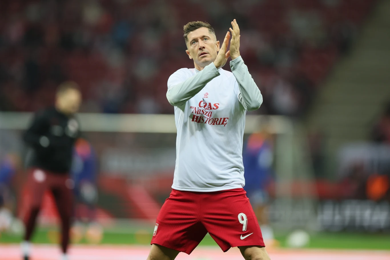 Piłkarz ubrany w białą koszulkę z napisem i czerwone spodenki stoi na boisku stadionowym, wykonując gest oklasków. W tle rozmazane sylwetki innych osób oraz trybuny stadionu.