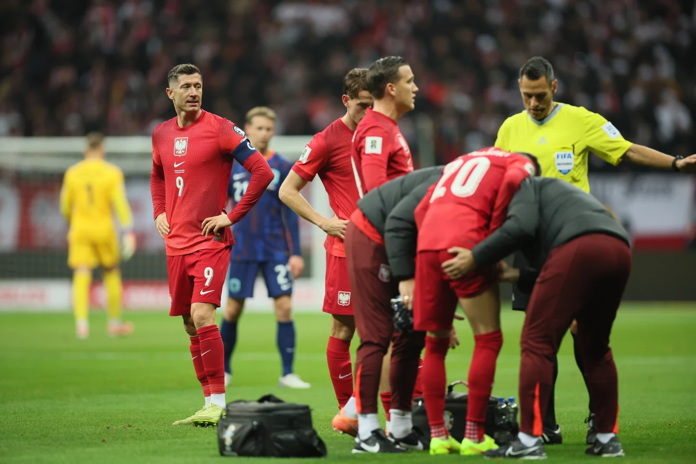 Jeszcze przed upływem kwadransa murawę musiał opuścić kontuzjowany Sebastian Szymański Polscy piłkarze w czerwonych strojach zgromadzeni na boisku podczas meczu, jeden z zawodników trzyma się za nogę i jest opatrywany przez członków sztabu medycznego, w tle widoczny sędzia oraz bramkarz w żółtym stroju.