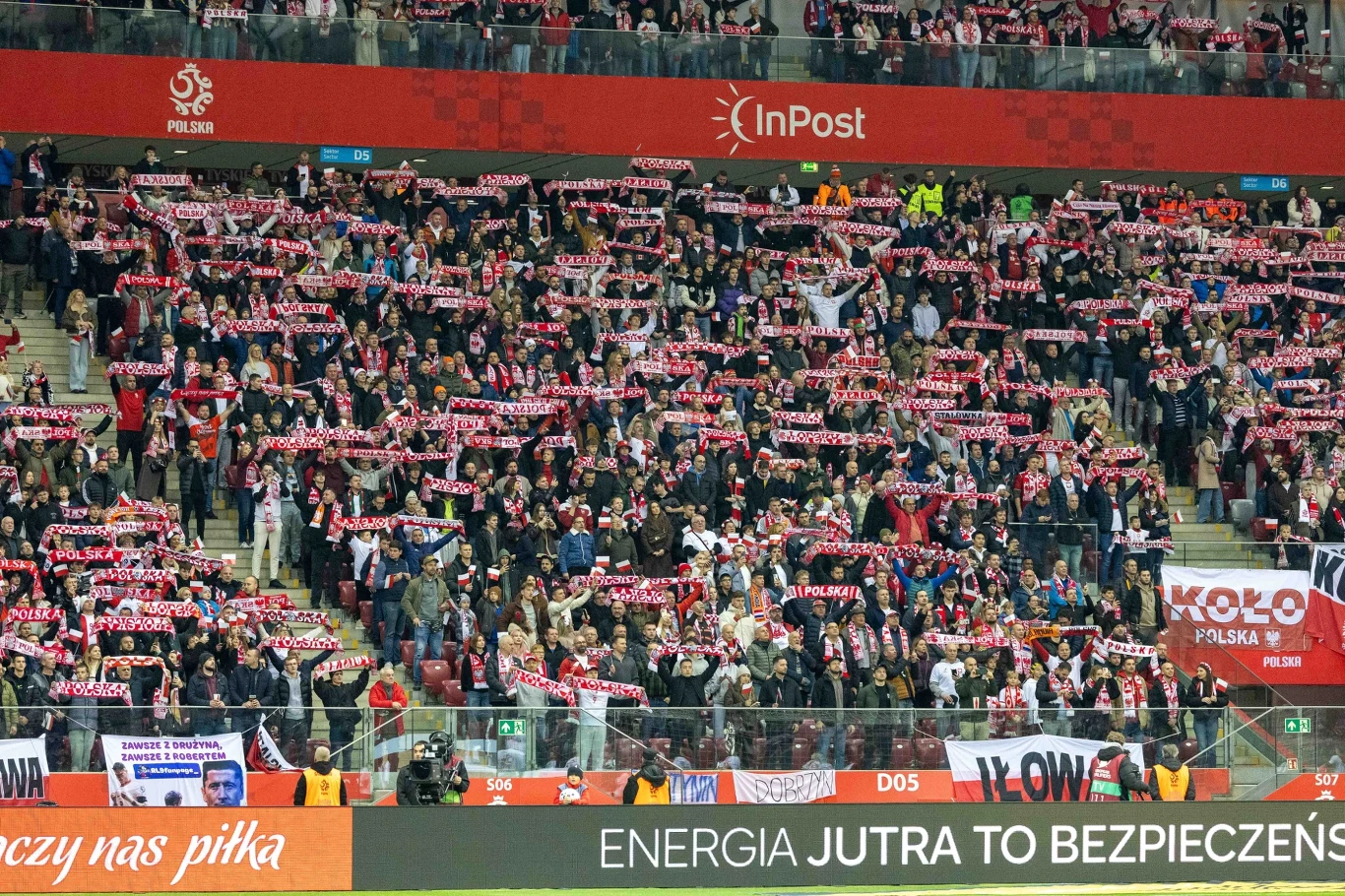 Kibice na Narodowym Tłum kibiców na trybunach stadionu piłkarskiego, większość osób trzyma czerwono-białe szaliki polskiej reprezentacji w górze. Banner InPost oraz inne reklamy i napisy widoczne na tle trybun.