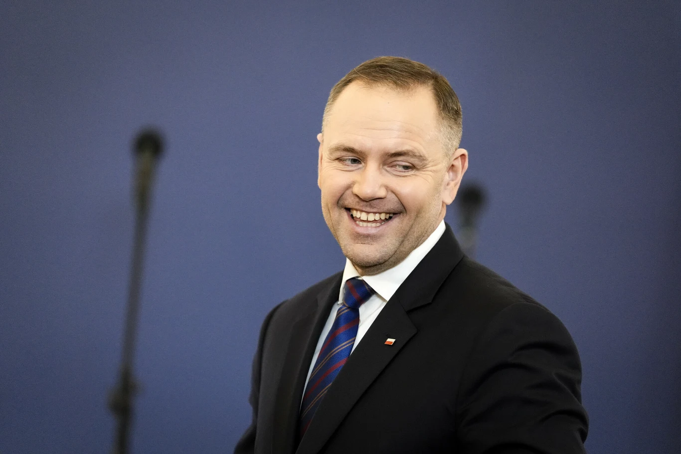 Karol Nawrocki Mężczyzna w garniturze z krawatem, uśmiecha się szeroko, tło jest jednolite, rozmyte, z mikrofonami ustawionymi w tle.