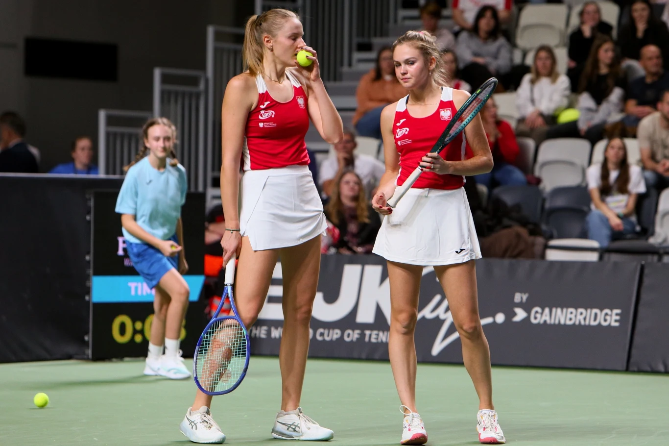 Dwie tenisistki w czerwonych koszulkach i białych spódniczkach stoją na korcie tenisowym, jedna trzyma rakietę i piłkę, druga opiera rakietę o nogę, w tle widownia oraz sędzina w niebieskim stroju.