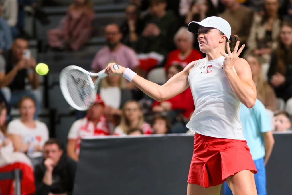 Tenisistka w białej czapce i stroju wykonuje dynamiczne uderzenie forhendowe na korcie, wokół zawodniczki widoczna jest publiczność uważnie obserwująca mecz.