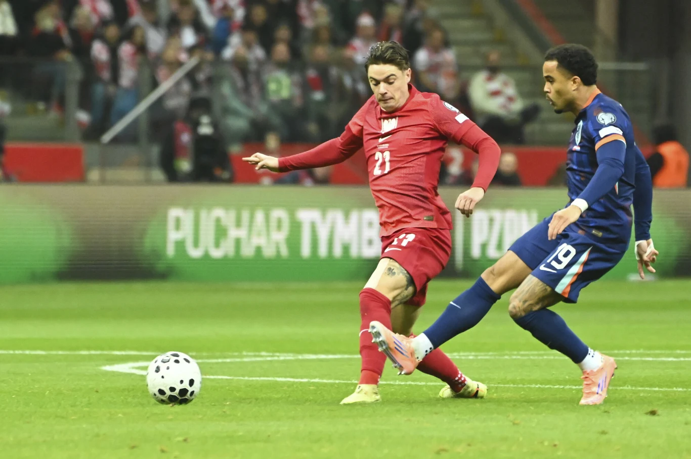 Dwóch piłkarzy rywalizuje o piłkę na boisku; jeden ubrany w czerwony strój, drugi w granatowy. W tle widoczna publiczność oraz reklamy przy boisku.