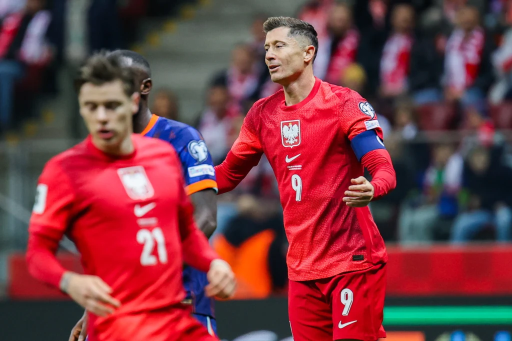 Nicola Zalewski i Robert Lewandowski w meczu Polska - Holandia Piłkarze w czerwonych strojach reprezentacji Polski podczas meczu piłkarskiego, jeden z zawodników z opaską kapitańską na ramieniu, na trybunach widoczna publiczność w biało-czerwonych barwach.