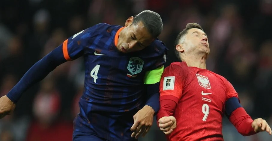 Dwóch piłkarzy walczy o piłkę głową podczas meczu; zawodnik w granatowej koszulce z numerem 4 i opaską kapitana zderza się z zawodnikiem w czerwonej koszulce z numerem 9.