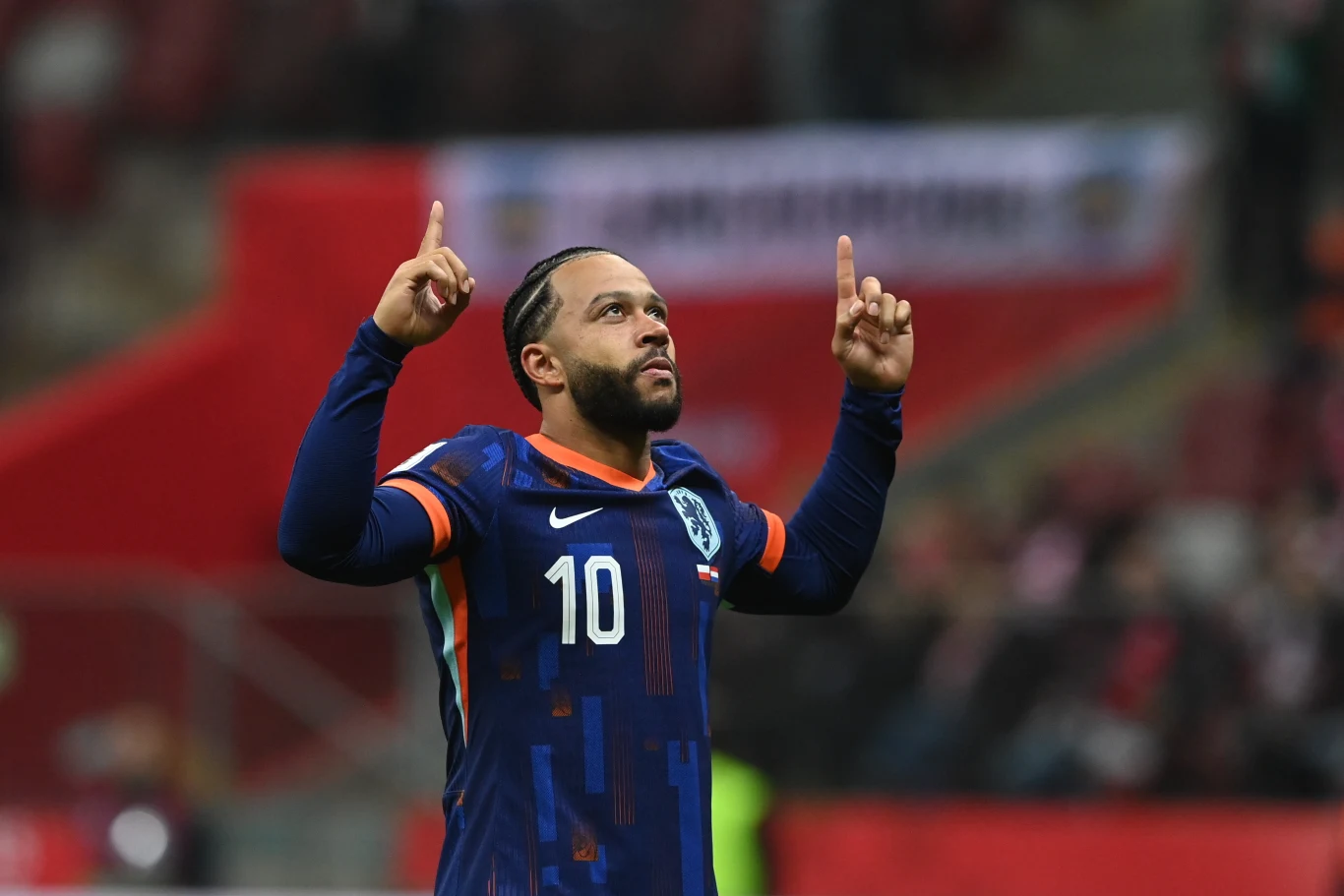 Piłkarz w granatowej koszulce z numerem 10 i reprezentacyjnymi barwami Holandii unosi obie ręce do góry, wskazując palcami niebo. W tle rozmazane trybuny stadionu oraz kibice.