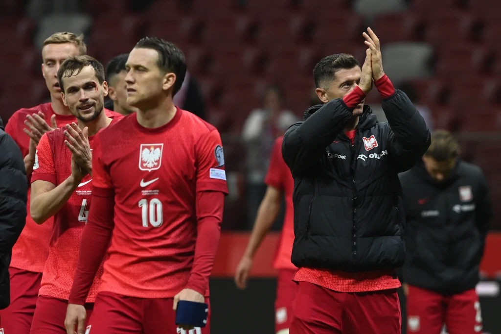 Polscy piłkarze w czerwonych strojach po zakończonym meczu na stadionie. Jeden z zawodników aplauzuje, a inni idą obok siebie. W tle trybuny, atmosfera sportowa.