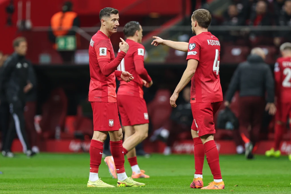 Piłkarze reprezentacji Polski w trakcie dyskusji na boisku podczas meczu, ubrani w czerwone stroje meczowe, w tle widać fragment stadionu i pojedynczych zawodników.