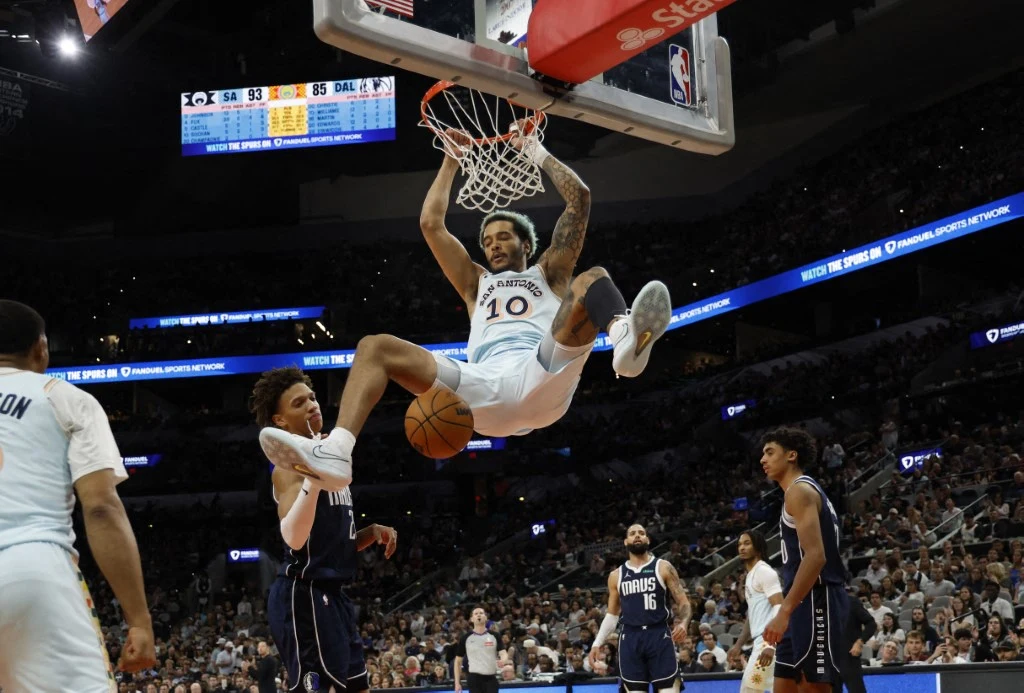 Zawodnik ligi NBA wykonuje efektowny wsad do kosza podczas meczu koszykówki, kilku innych graczy obserwuje akcję pod koszem na tle wypełnionych trybun.