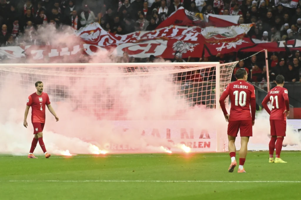 Zawodnicy w czerwonych strojach stoją na murawie obok bramki, wokół której unoszą się kłęby dymu z rac świetlnych, w tle widać tłum kibiców oraz duże biało-czerwone flagi kibicowskie.