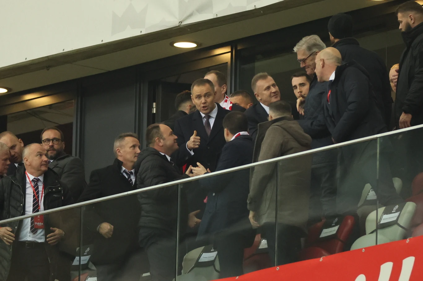 Karol Nawrocki zabrał głos po meczu Polska - Holandia Grupa mężczyzn w garniturach zgromadzona na trybunie stadionu, część z nich prowadzi rozmowy i wymienia gesty, jeden z mężczyzn pokazuje kciuk uniesiony w górę.