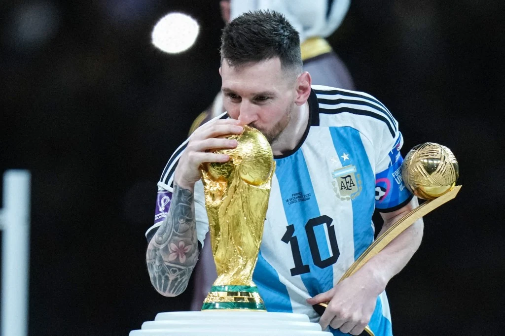 Piłkarz w koszulce reprezentacji Argentyny całuje Złotą Kulę FIFA, trzymając statuetkę Złotego Buta, świętując zwycięstwo w turnieju piłkarskim.