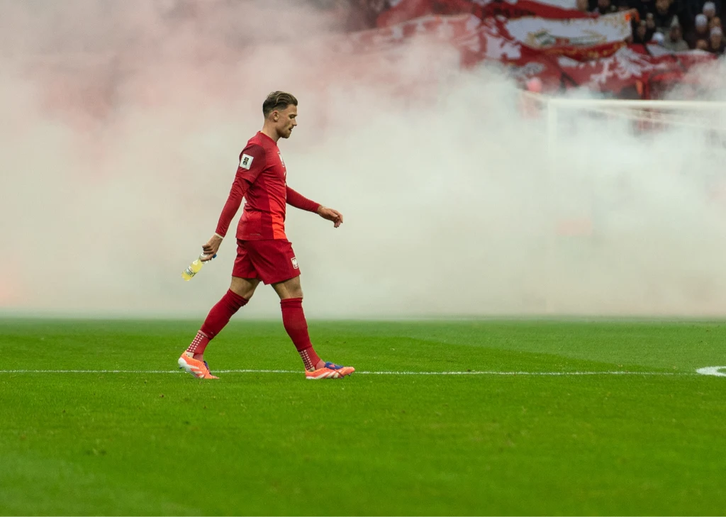 Matty Cash w meczu Polska - Holandia Piłkarz ubrany w czerwony strój sportowy idzie po murawie boiska z wyrzutnią dymu w ręku, w tle rozprzestrzenia się gęsty biały dym, a trybuny są częściowo zasłonięte.