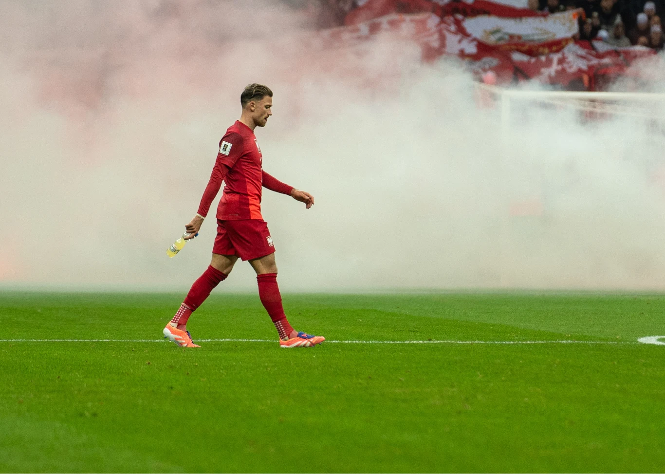 Piłkarz ubrany w czerwony strój sportowy idzie po murawie boiska z wyrzutnią dymu w ręku, w tle rozprzestrzenia się gęsty biały dym, a trybuny są częściowo zasłonięte.