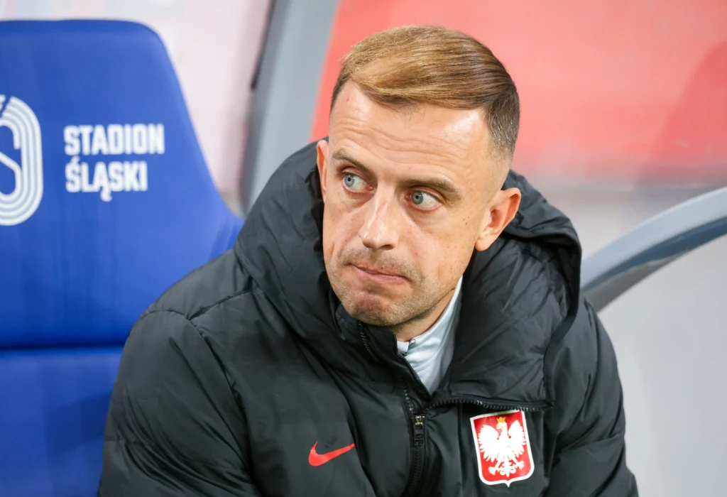 Kamil Grosicki Mężczyzna w kurtce sportowej z herbem Polski, siedzący na ławce przy stadionie, w tle widoczny fragment napisu STADION ŚLĄSKI i niebieskie oparcie.