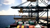 Port przeładunkowy z kontenerowcem cumującym przy nabrzeżu, widoczne dźwigi załadowcze unoszą kontenery, w tle widać magazyny i morze, podkreślające skalę działalności logistycznej.