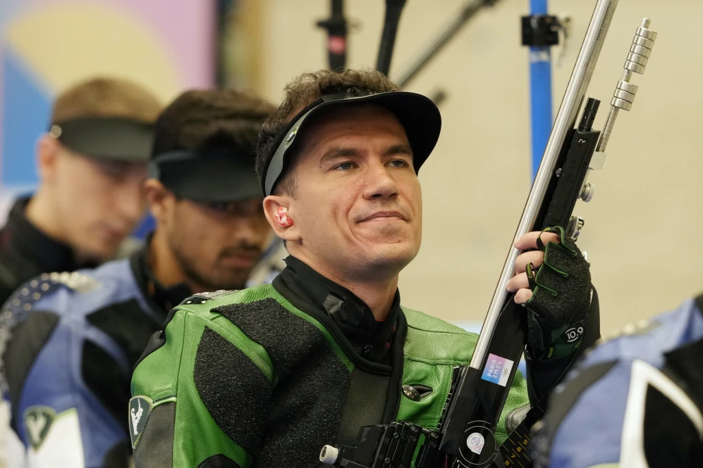 Mężczyzna w stroju sportowym do strzelectwa trzyma karabin, w tle kilku innych zawodników skupionych na swoich zadaniach, cała scena odbywa się w zamkniętej hali sportowej.