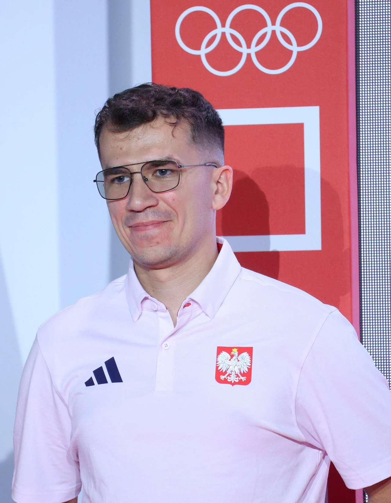 Mężczyzna w okularach w białej koszulce z godłem Polski oraz logo sportowym stoi na tle czerwonej tablicy z symbolami olimpijskimi.