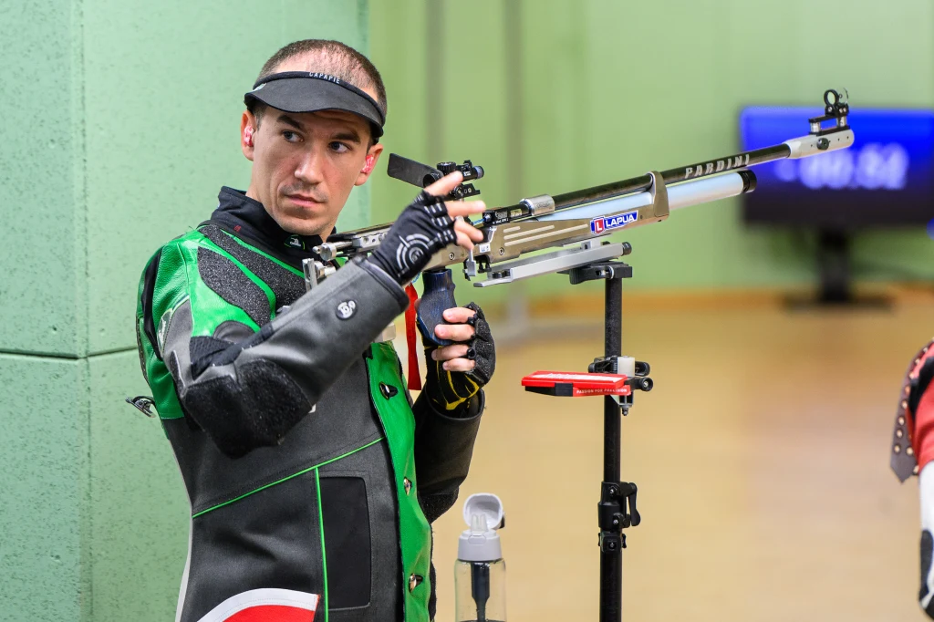 Zawodnik w stroju ochronnym i rękawicach celuje z karabinu sportowego w kierunku tarczy na profesjonalnej strzelnicy. W tle zielona ściana i rozmyty wyświetlacz elektroniczny.