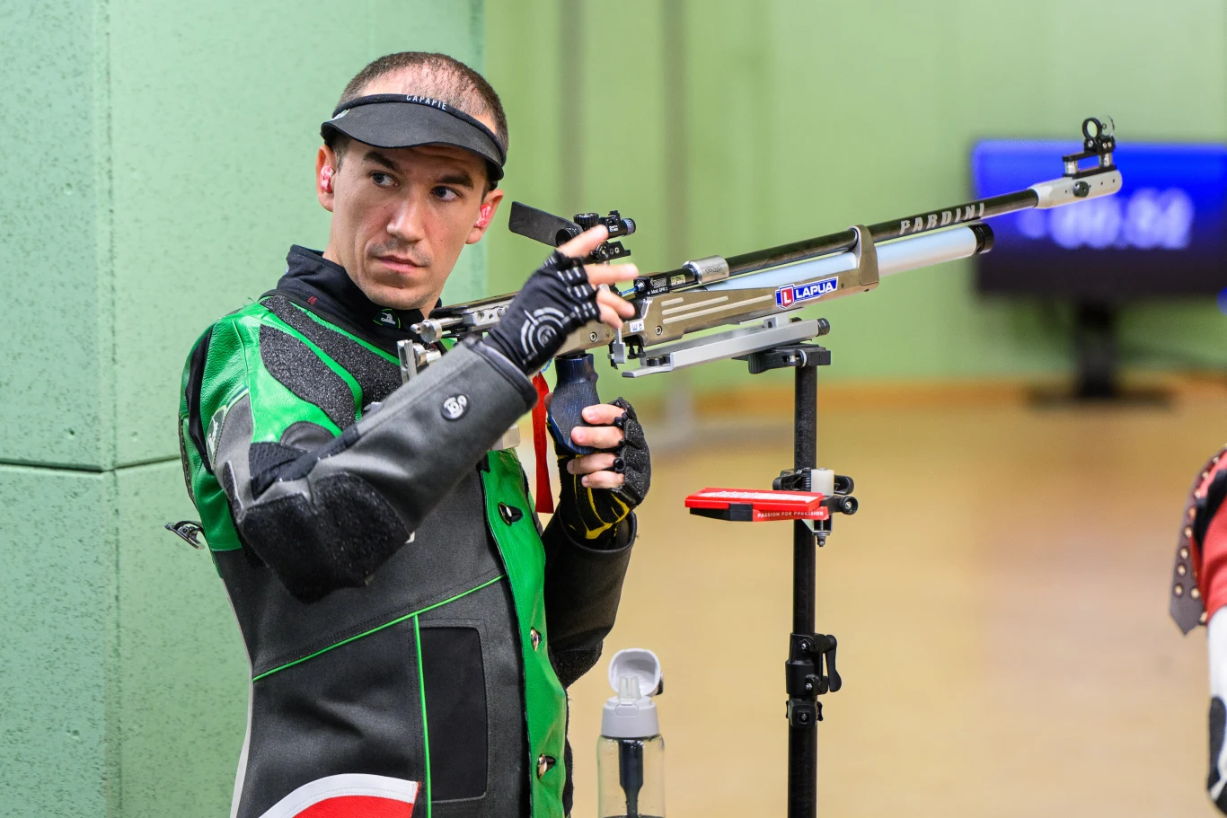 Zawodnik w stroju ochronnym i rękawicach celuje z karabinu sportowego w kierunku tarczy na profesjonalnej strzelnicy. W tle zielona ściana i rozmyty wyświetlacz elektroniczny.