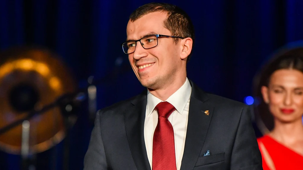 Uśmiechnięty mężczyzna w garniturze i czerwonym krawacie stoi na scenie na tle niebieskiego oświetlenia, obok niego kobieta w czerwonej sukni z upiętymi włosami.