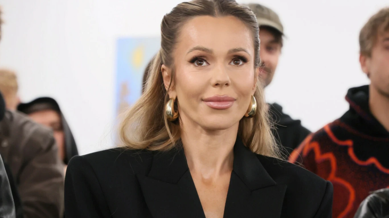 Doda Kobieta o długich blond włosach spiętych do tyłu, ubrana w elegancki czarny żakiet i duże złote kolczyki. W tle widoczne są inne osoby, nieostro określone, co sugeruje obecność na wydarzeniu lub spotkaniu.