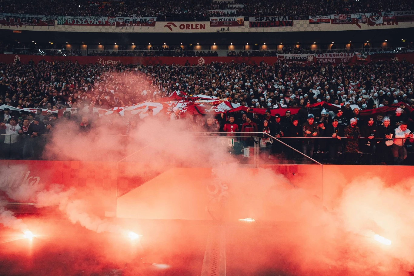 Kibice podczas meczu Polska - Holandia rzucali racami Tłum kibiców na stadionie z rozciągniętą flagą narodową, unoszący się gęsty dym i światło rac tworzą intensywną atmosferę podczas wydarzenia sportowego.