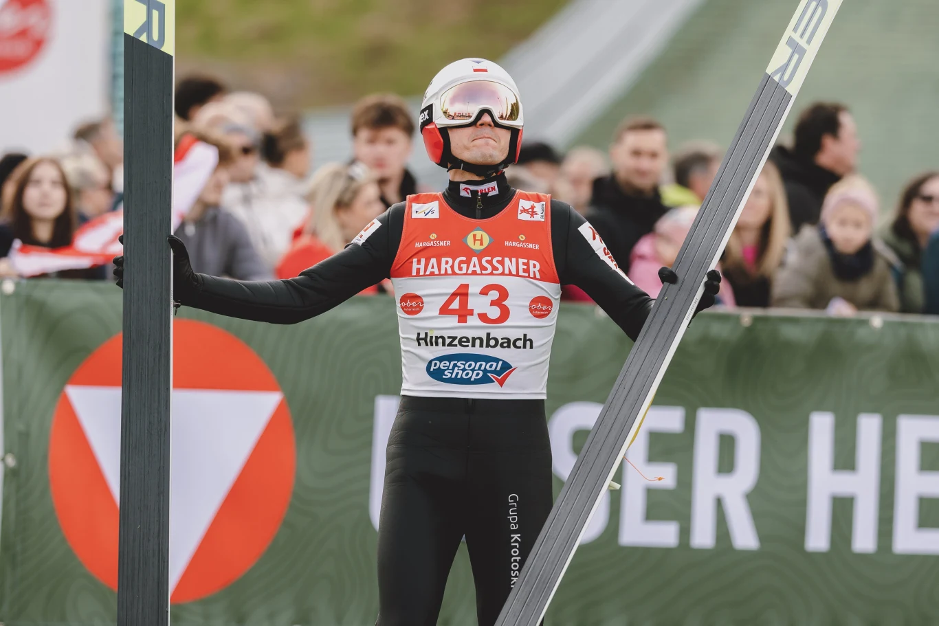 Kamil Stoch Zawodnik skoków narciarskich w kasku i goglach stoi na tle tłumu kibiców, ubrany w specjalny kombinezon i trzyma narty w obu rękach, na piersi widoczny numer startowy 43 oraz logo sponsorów.