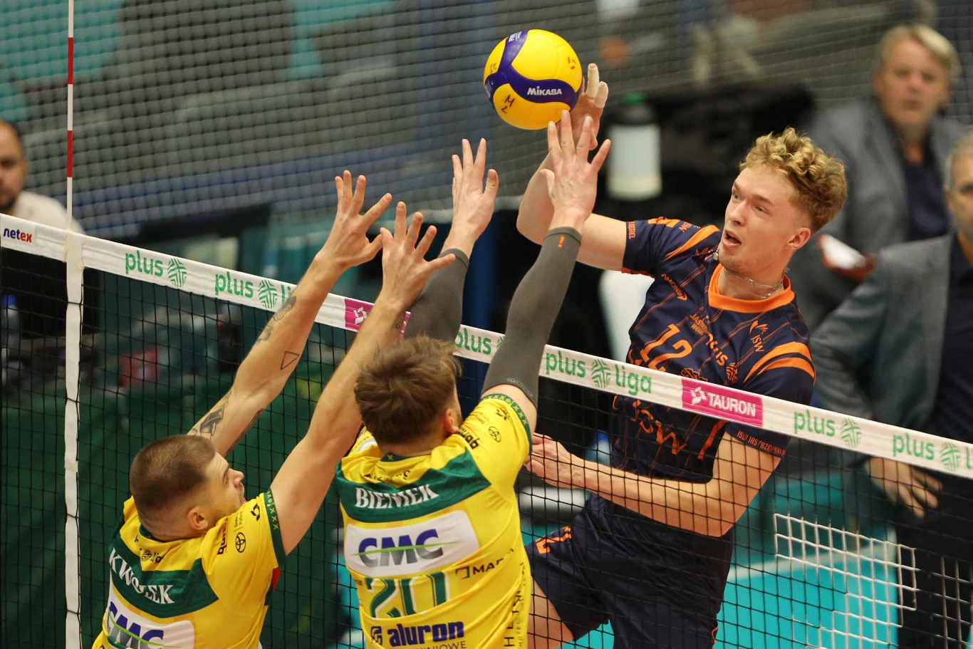 Zawodnik w pomarańczowo-granatowym stroju wykonuje atak na piłkę podczas meczu siatkówki, podczas gdy dwóch graczy w żółto-zielonych koszulkach próbuje zablokować ruch przy siatce.