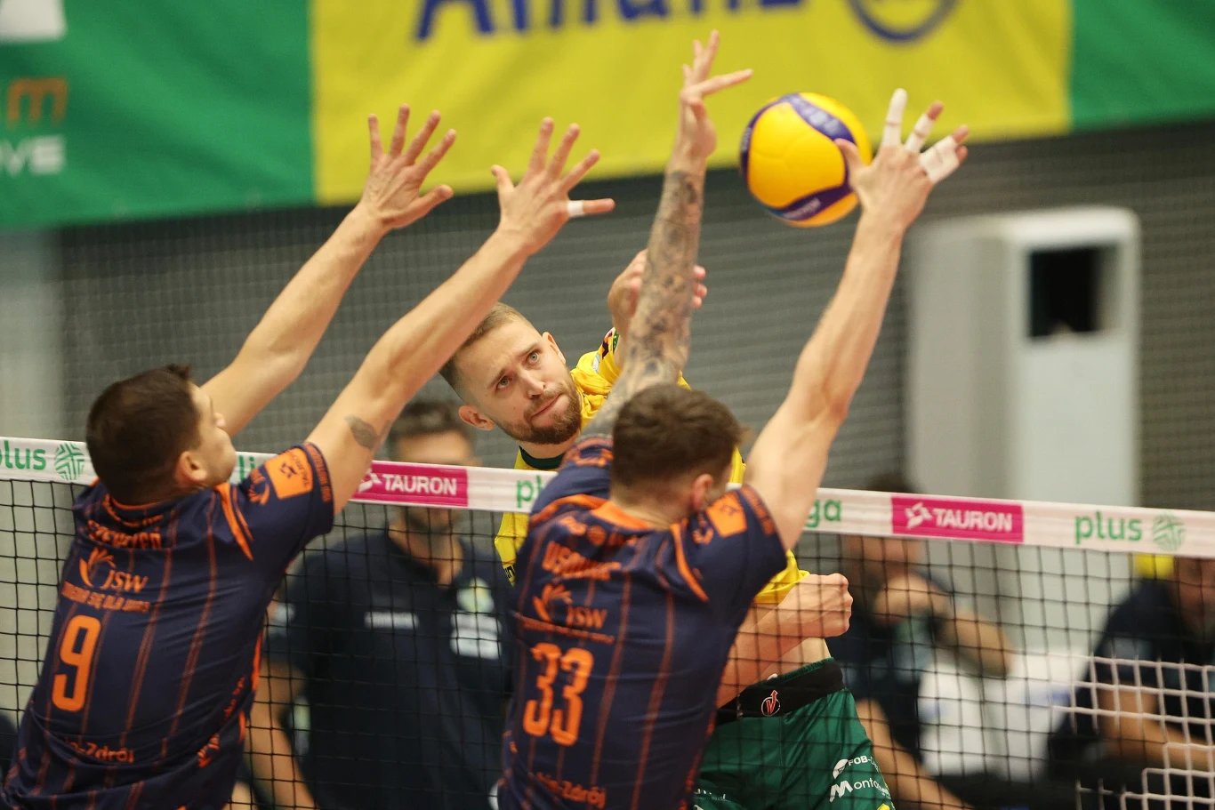 Zawodnik w żółto-zielonym stroju wykonuje atak piłką podczas meczu siatkówki, dwóch blokujących w ciemnych koszulkach z numerami 9 i 33 próbuje zatrzymać piłkę na siatce, w tle widać fragment trybun oraz reklamowe banery.